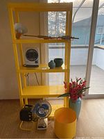 Yellow plastic shelf unit with white portable heater, two ceramic pots (teal and dark green), yellow planter, large blue vase with red artificial flowers at the base, yellow desk lamp, black portable fan, and blue metal fan.
