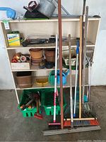 Front view of shelving unit showing pots, watering cans, broom, rakes, and other garden tools leaning against unit.