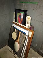 Stack of various framed pictures including a print with wooden spoons in front seen against a concrete wall
