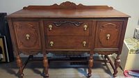 Front view of antique wooden buffet showing detailed carvings, two drawers in center, one cabinet door on each side, brass handles, and turned legs.