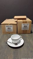 Two sealed brown boxes with product labels, plus one opened set showing one white ceramic cappuccino cup, saucer, and silver-colored spoon on a wooden surface.