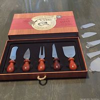 Overall view of five piece Petit Maison cheese spreader set in original box with wooden handles and metal blades, displayed with five plastic blade covers on the side for protection.