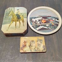 Three vintage decorative tins on table: horse riding woman, Expo 67 round tin, small kittens tin.