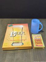 Box of The Beer Game board game, Shot Recipe Jumbo Playing Cards deck, and blue plastic freezer mug arranged side by side.
