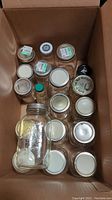 Top-down view of 14 assorted clear glass jars, including 12 large 1-liter jars with various lids in white, black, and metallic colors, arranged inside a cardboard box.