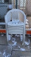 Stack of six white plastic lawn chairs outdoors on concrete with chain-link fence background.