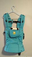 Front view of teal Lille baby carrier hanging on white plastic hanger against beige wall. Features padded shoulder straps, black buckles, front pouch with zipper, snap buttons near base.