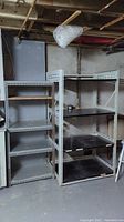 Photo showing two metal shelving units. The smaller unit has wooden shelves and is narrower and shorter. The larger unit has black metal shelves and is deeper and taller. Both have perforated metal supports.