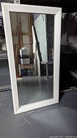 White framed rectangular mirror standing upright against shelves in storage area.