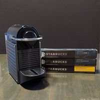 Black Nespresso Pixie C60 coffee machine with 3 sleeves of Starbucks coffee capsules beside it on a dark surface.