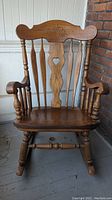 Full view of solid oak rocking chair showing carved floral backrest with heart cutout, spindle arms and legs, and curved rockers.