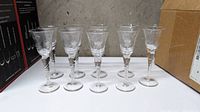 Nine ornate etched clear glass shot glasses with twisted stems, arranged in two rows on a white surface with boxes in background.