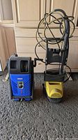 Photo shows two electric power washers side by side on a concrete floor in front of wooden cabinetry. The left unit is blue and black, the right unit is yellow and black with hose and power cord coiled on it.