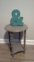Wooden round side table with four turned legs and a lower triangular shelf, topped with a turquoise ampersand decorative piece.