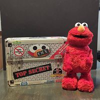 Photo of Tenth Anniversary Tickle Me Elmo plush and its themed metal carrying case, showing details of the packaging and toy.