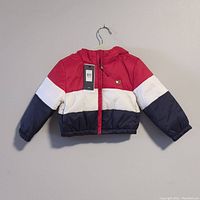 Front view of the Tommy Hilfiger color-block jacket hanging on a hanger, showing red, white, and navy blue sections with a hood and a tag still attached.