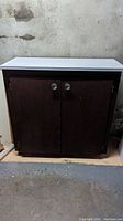 Two-door lower cabinet with a white countertop positioned against a concrete wall, dark brown finish, round metal knobs on doors