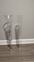 Photo of two tall glass vases against a gray wall on a wood floor. One vase has a spiral design and flared top, the other is plain and cylindrical.
