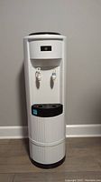 Front view of white and black water cooler standing against a gray wall on wood flooring.