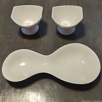 Two white serving bowls with pedestal bases and a white double-bowl serving dish in a figure-eight shape on dark surface.