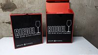 Two Riedel branded boxes, one open and one closed sitting on a flat surface with a concrete wall background.