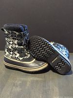 Black and white patterned women's Sorel winter boots, one boot standing upright and the other showing the sole with tread and Sorel branding.