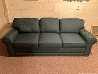 Front view of green upholstered three-seat couch with three cushions and patterned armrests, placed against a beige brick wall on brown carpet.