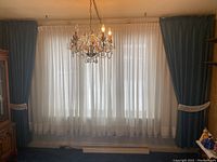 Full view of window with two sets of curtains: sheer white inner layer and blue outer drapes with tassel tiebacks hanging on either side, stained and requiring cleaning.