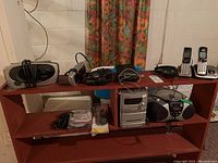 Photo of a shelf with various electronic devices including clock radios, a CD player with radio, cordless phones with base units, and earphones.