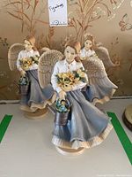 Photo showing three identical winged female angel figurines standing together on a shelf, each holding yellow flowers and a watering can