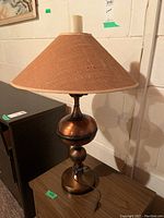 Full view of the metal base lamp with woven fabric lampshade, sitting on a wooden surface against a white wall.