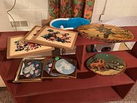 Photo showing seven framed floral and nature prints and two oval laminated wood block prints placed on wooden shelves in a basement setting.