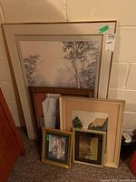 Photo showing all five framed prints stacked against a wall in basement storage area, varying sizes and frame styles visible.