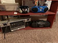 Four vintage portable radio and cassette players displayed on a red shelf on a carpeted floor.
