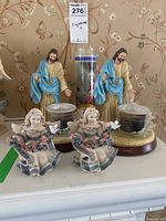 Front view of two Jesus figurines in beige and blue robes with candle holders, two matching angel figurines holding white doves surrounded by floral garlands, and a tall religious candle in the background.