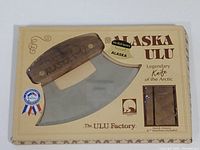 Photo of the Alaska Ulu knife in its original packaging, showing the wooden handle with Alaska printed on it, curved stainless steel blade, and product branding.