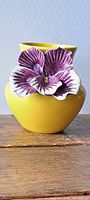 Close-up photo of a yellow ceramic vase with a large purple and white flower attached to its side, showing the front view of the item.