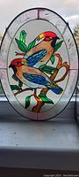 Front view of the oval hand painted glass showing two birds perched on a leafy branch with chain hanger.