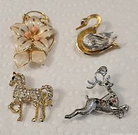 Lot of 4 brooches displayed together on a white background: white flower with pink beads, gold and silver swan, rhinestone-covered gold horse, and silver-tone reindeer with rhinestones.