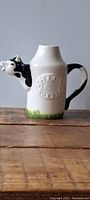 Photo of the cow-shaped ceramic creamer pitcher from the side, showing details of the cow head spout and grass decoration at base.