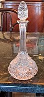 Tall cut crystal decanter with an intricate cut crystal stopper sitting on a marble surface, backdropped by wooden furniture.