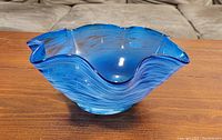 Side angled view of the decorative blue glass bowl, showing ruffled and wavy edge with translucent blue and darker striation details.
