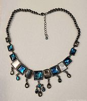 Photo shows a silver-tone chain necklace with square decorative pieces in blue, grey, and white shades, and teardrop-shaped pendants hanging along the bottom edge. Necklace measurement approx 17 inches.