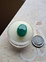 Photo of a large oval cut loose emerald stone displayed in a clear round plastic container, placed on a light-colored textured surface, next to a Canadian 10-cent coin for size reference.