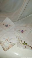 Four antique white hand embroidered hankies laid out, folded showing delicate embroidery and lace details.
