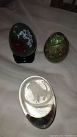 Three decorative eggs shown: two painted decorative eggs with floral and bird motifs, one mounted on a stand, and one solid clear glass egg with an etched lion paperweight design