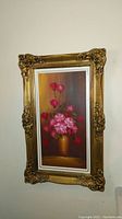 Full view of the framed oil painting showing pink roses in gold vase with ornate gold frame on a wall.