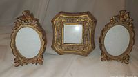 Three small decorative picture frames photographed together on a neutral background: two oval frames with bird and ribbon ornaments and one square frame with floral decorations.