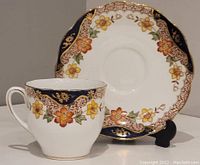 Cup and saucer set showing floral, gold, and navy blue Imari pattern design, displayed on black stand.