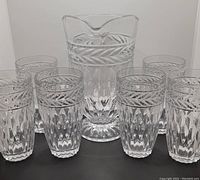 Front view of the footed pitcher centered among six matching glasses, showing the leaf and geometric cut pattern details.